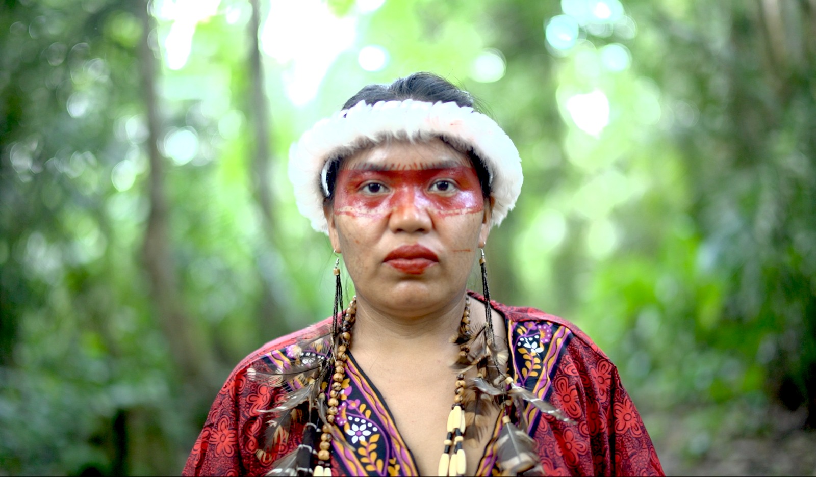 Woman with red face paint across her face, feathered earrings and a red top stands infront of a blurred forest background