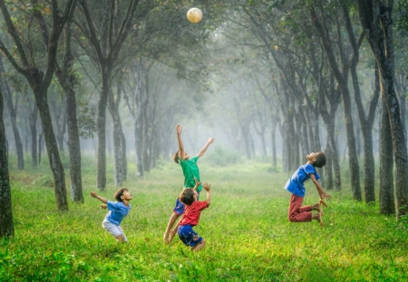 Four children jumping up to catch a football. They are playing on grass inbetween two tall rows of trees